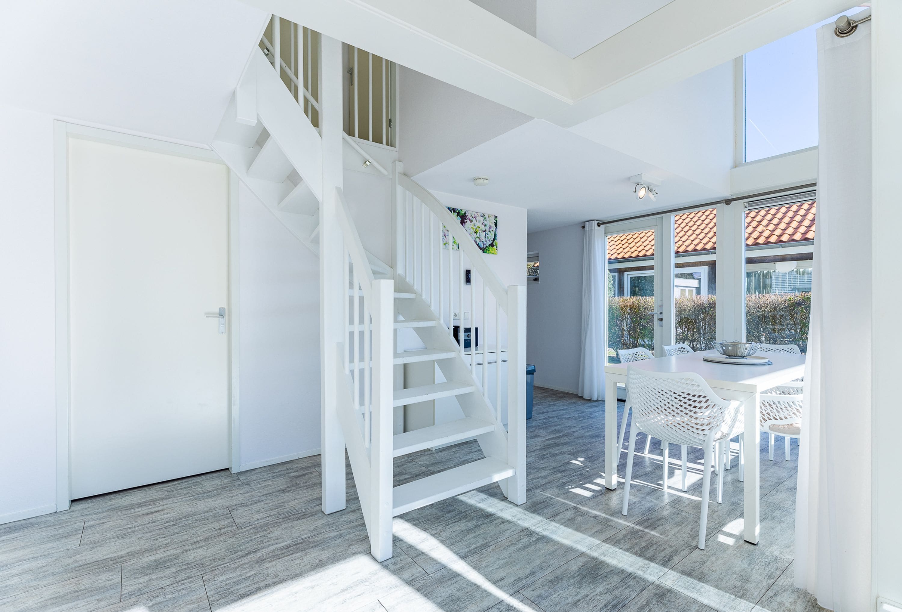 Un intérieur moderne avec un escalier blanc, un coin repas avec des meubles blancs et de grandes fenêtres.