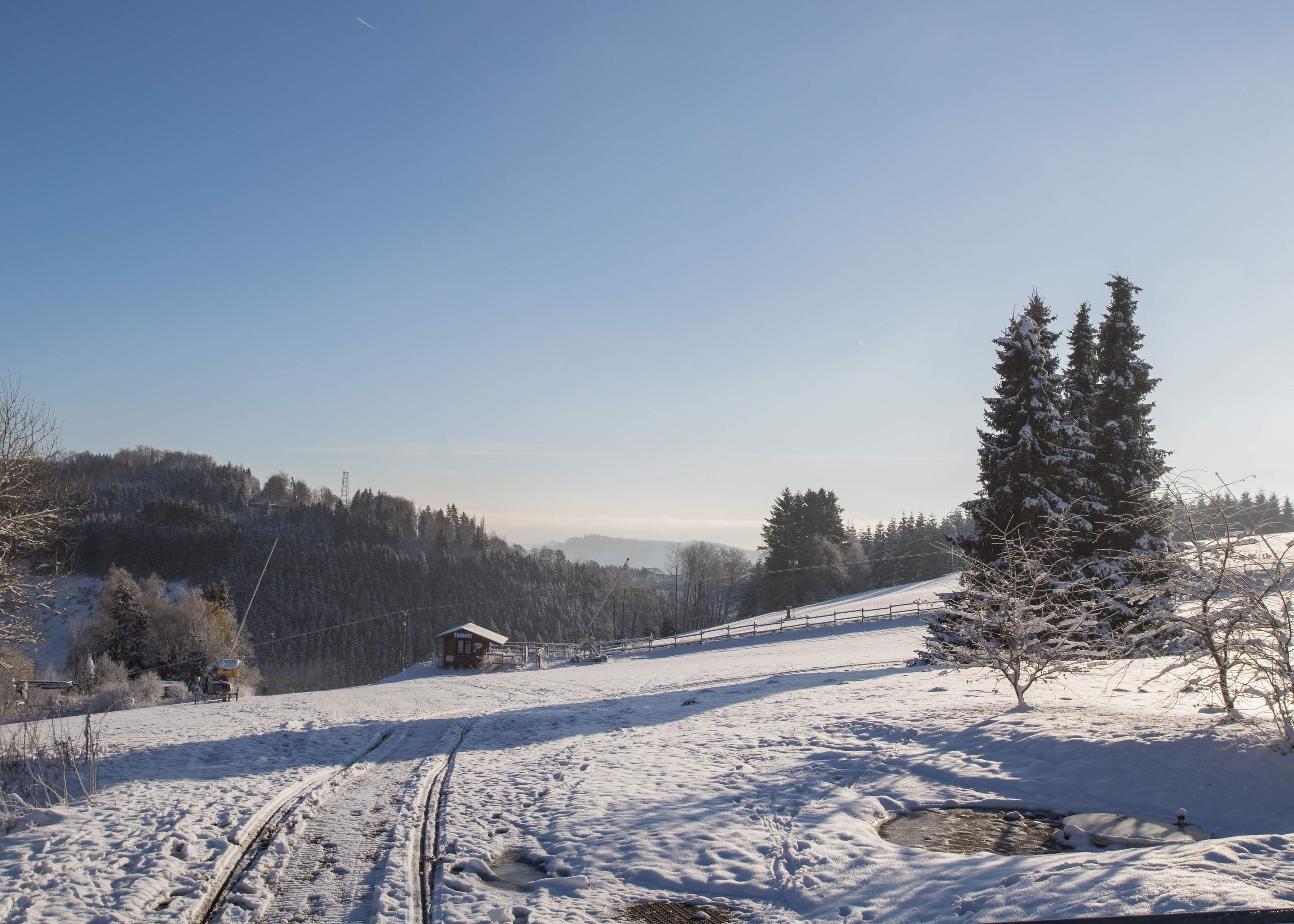Schneebedeckte Landschaft mit einer Holzhütte, blauem Himmel und einem bewaldeten Hügel.