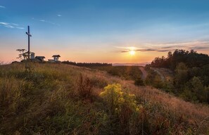Ein Kreuz, eine kleine Struktur und ein Pfad in einer grünen Landschaft, im Hintergrund ein Sonnenuntergang.