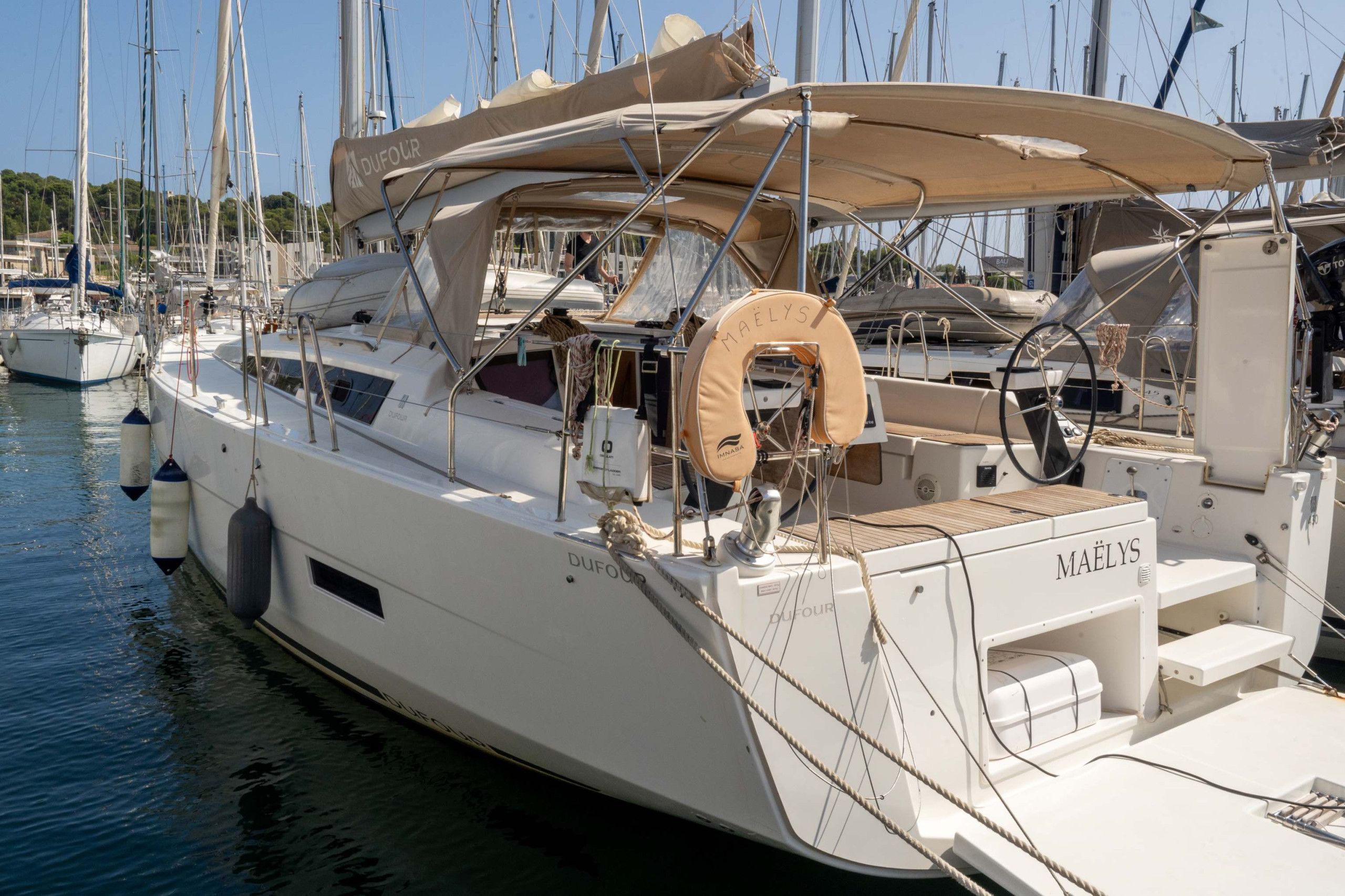 Un yacht blanc est amarré dans une marina, avec plusieurs mâts visibles en arrière-plan.