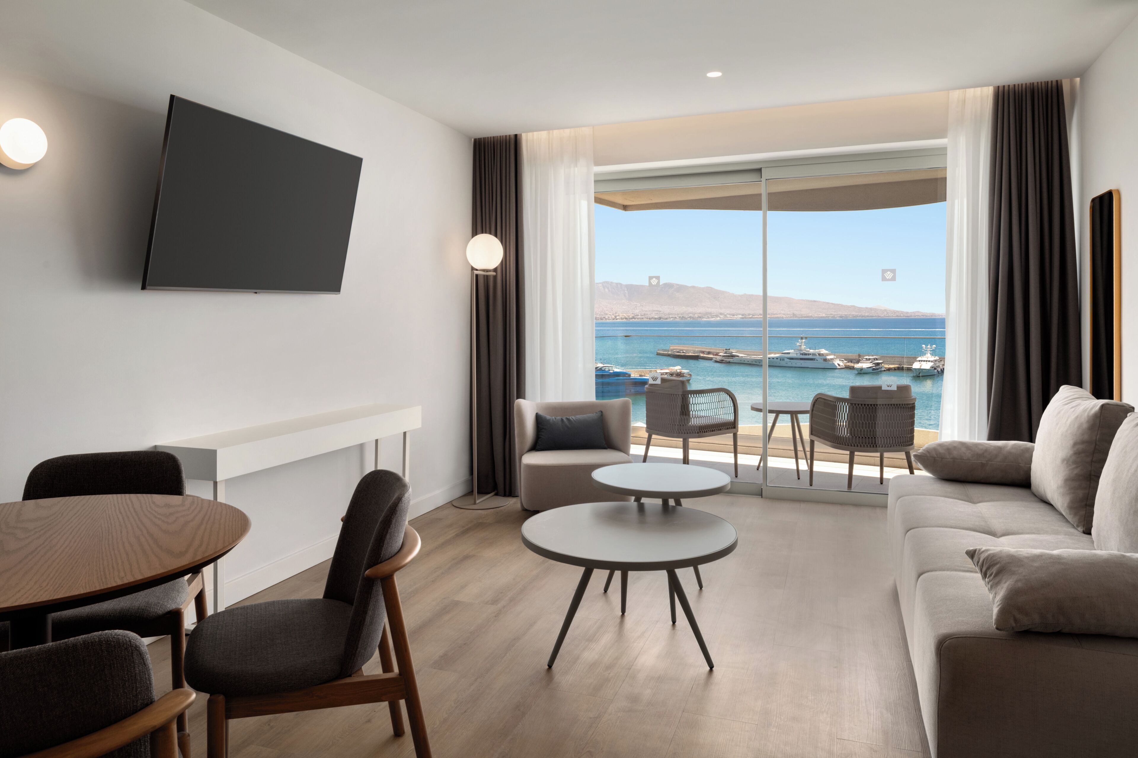 Quarto de hotel moderno com televisão de tela plana, área de refeições, sofá e vista para o mar e barcos.