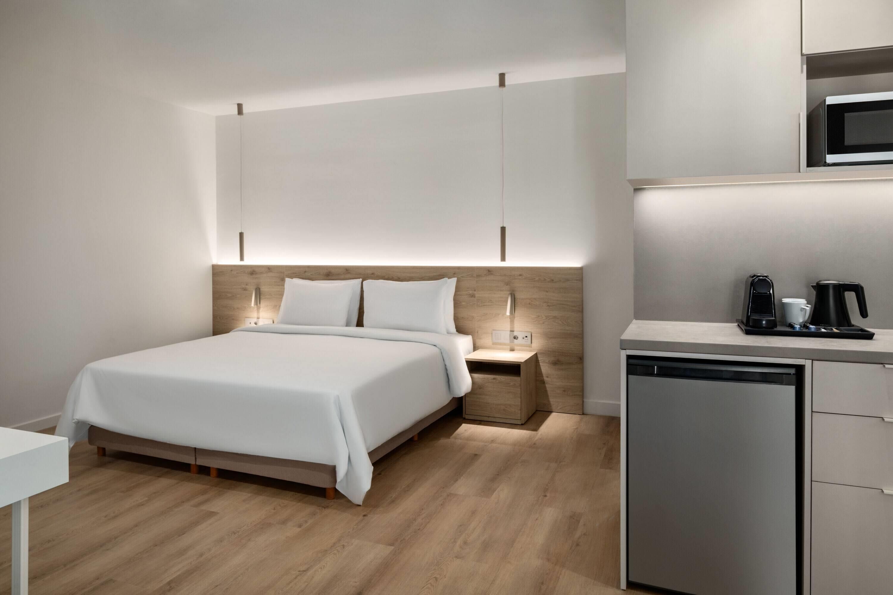 A modern hotel room with a bed, bedside table, and a built-in kitchen area.