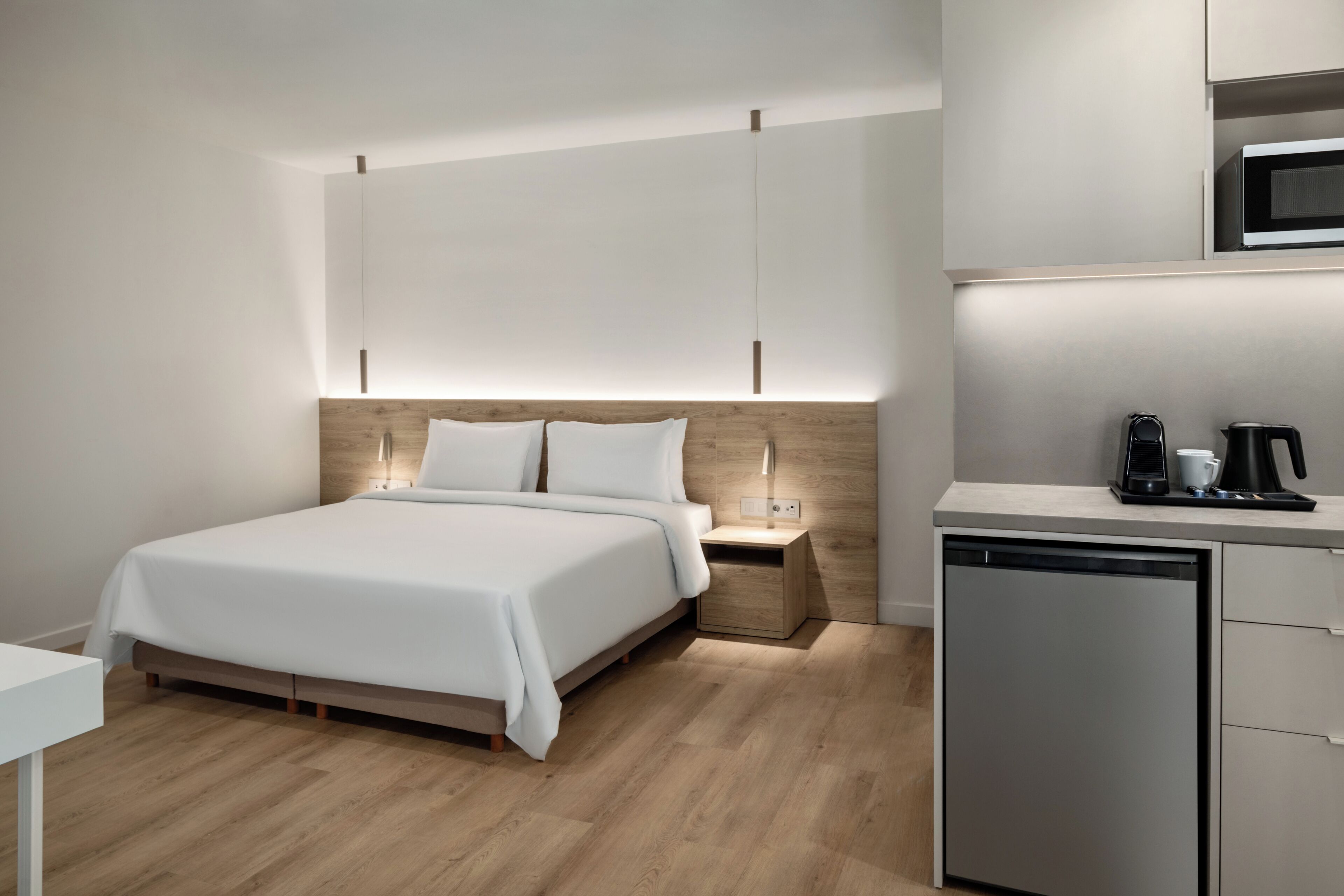 Una habitación de hotel moderna con cama, mesita de noche y una cocina integrada con microondas.