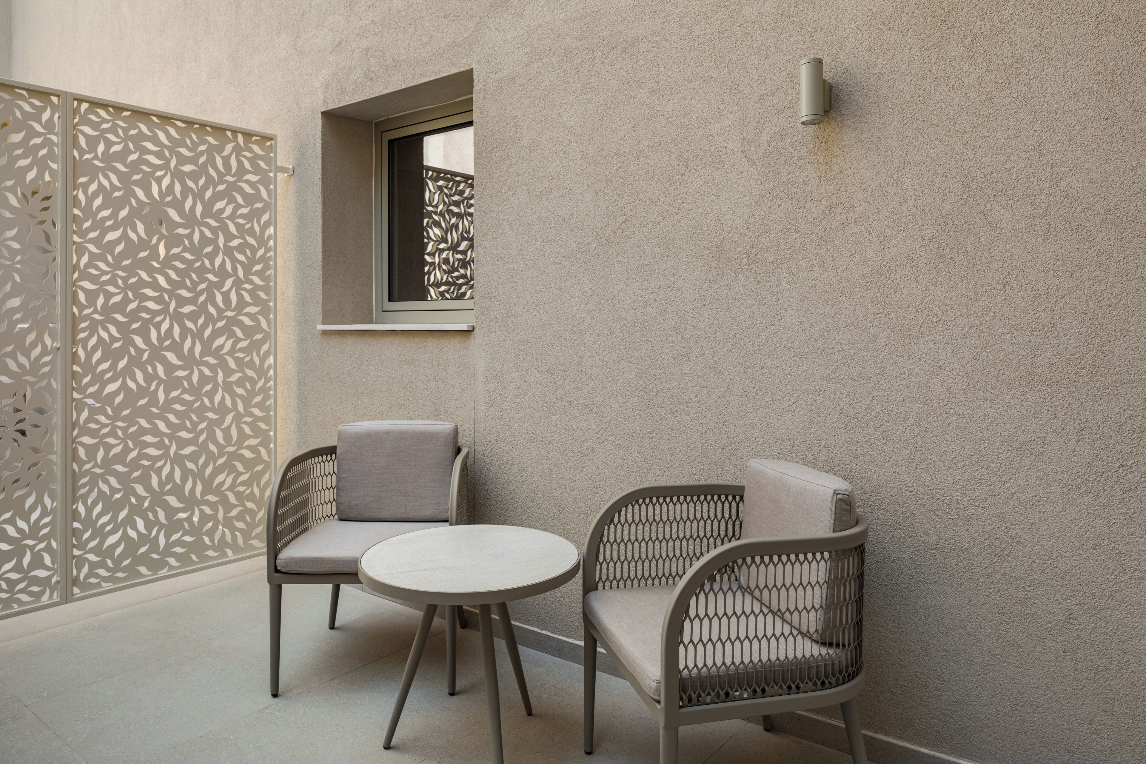 A small outdoor seating area with two chairs and a round table, a decorative wall with a leaf pattern, and a window with a view.