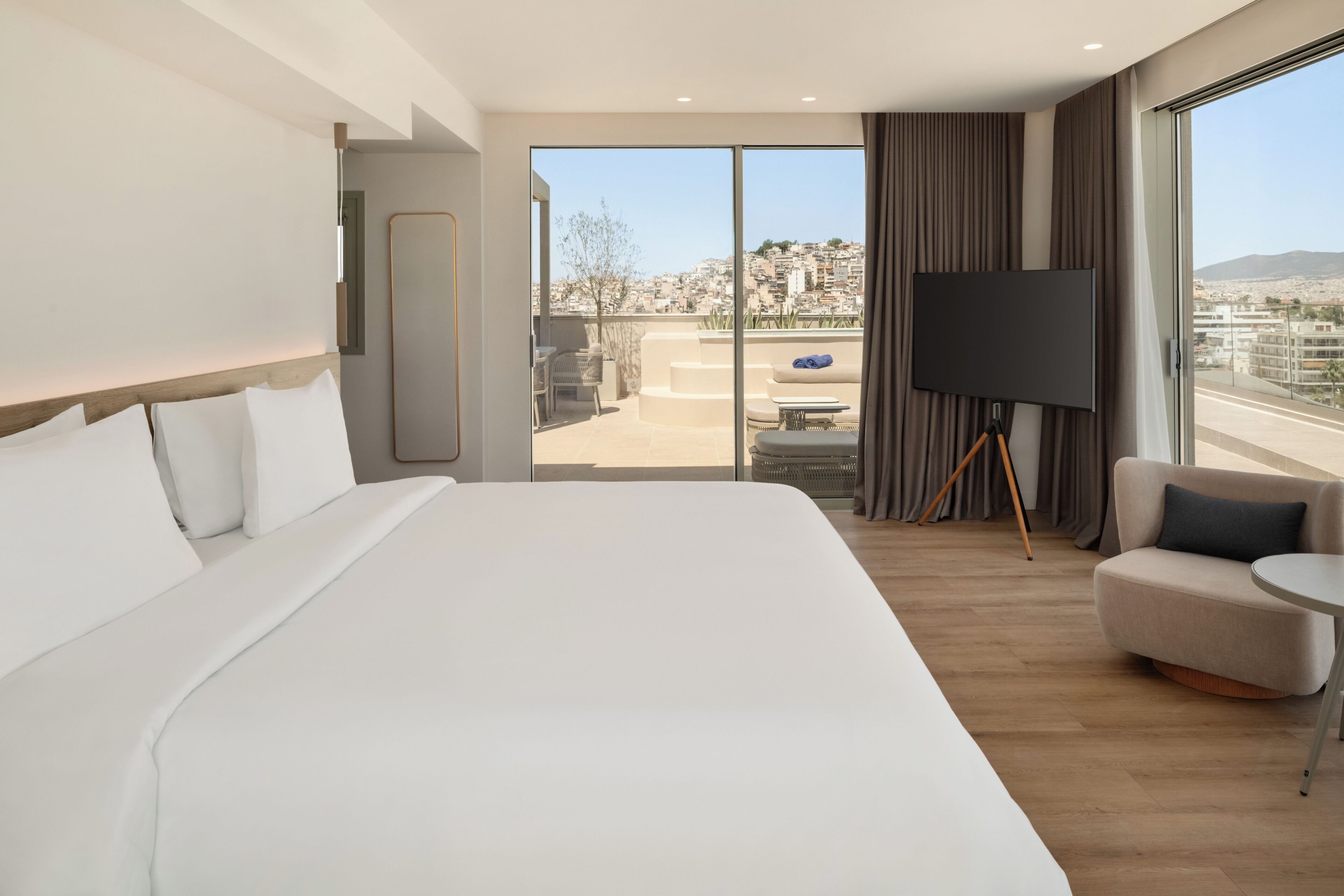 A modern hotel room with a large bed, a seating area with a TV, and a balcony with a city view.