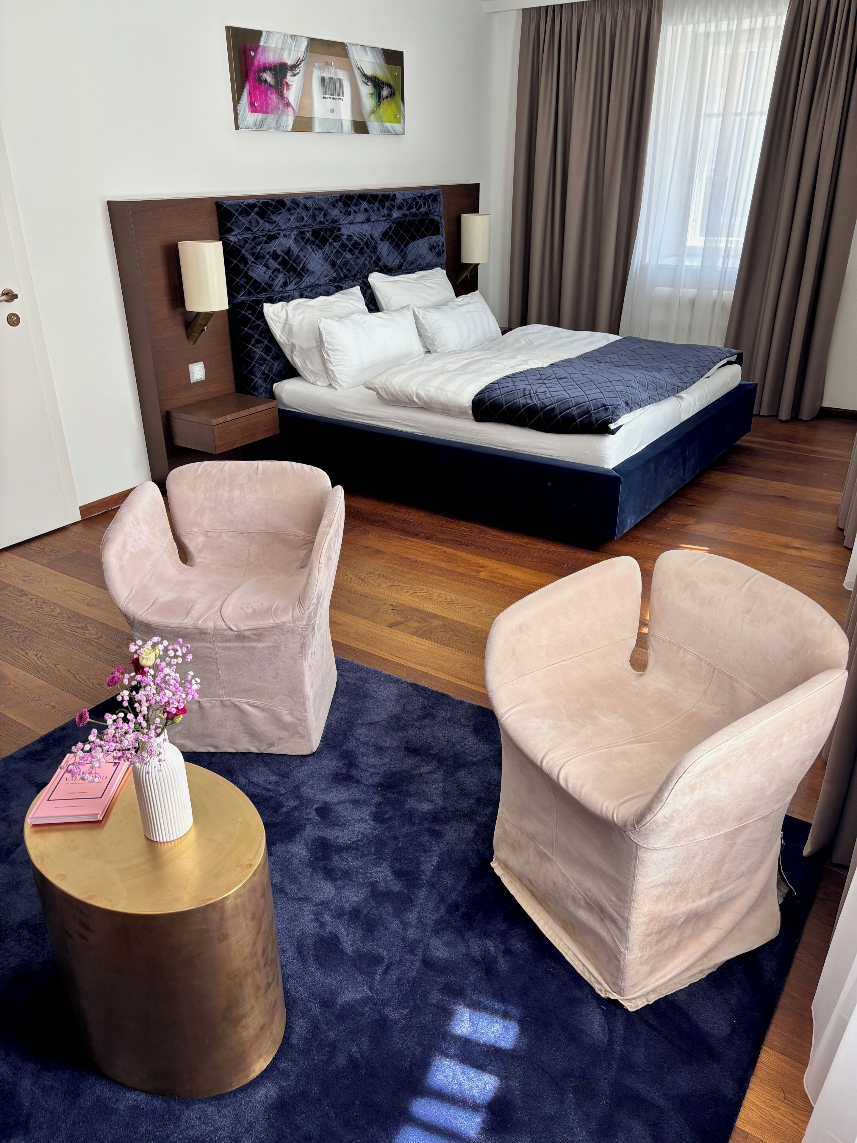 A hotel room with a bed, two armchairs, a small round table with a vase of flowers, and a painting on the wall.