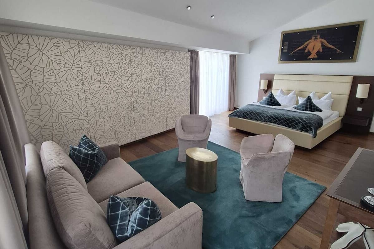 A modern hotel room with a sofa, armchair, round coffee table, and a bed with a headboard.