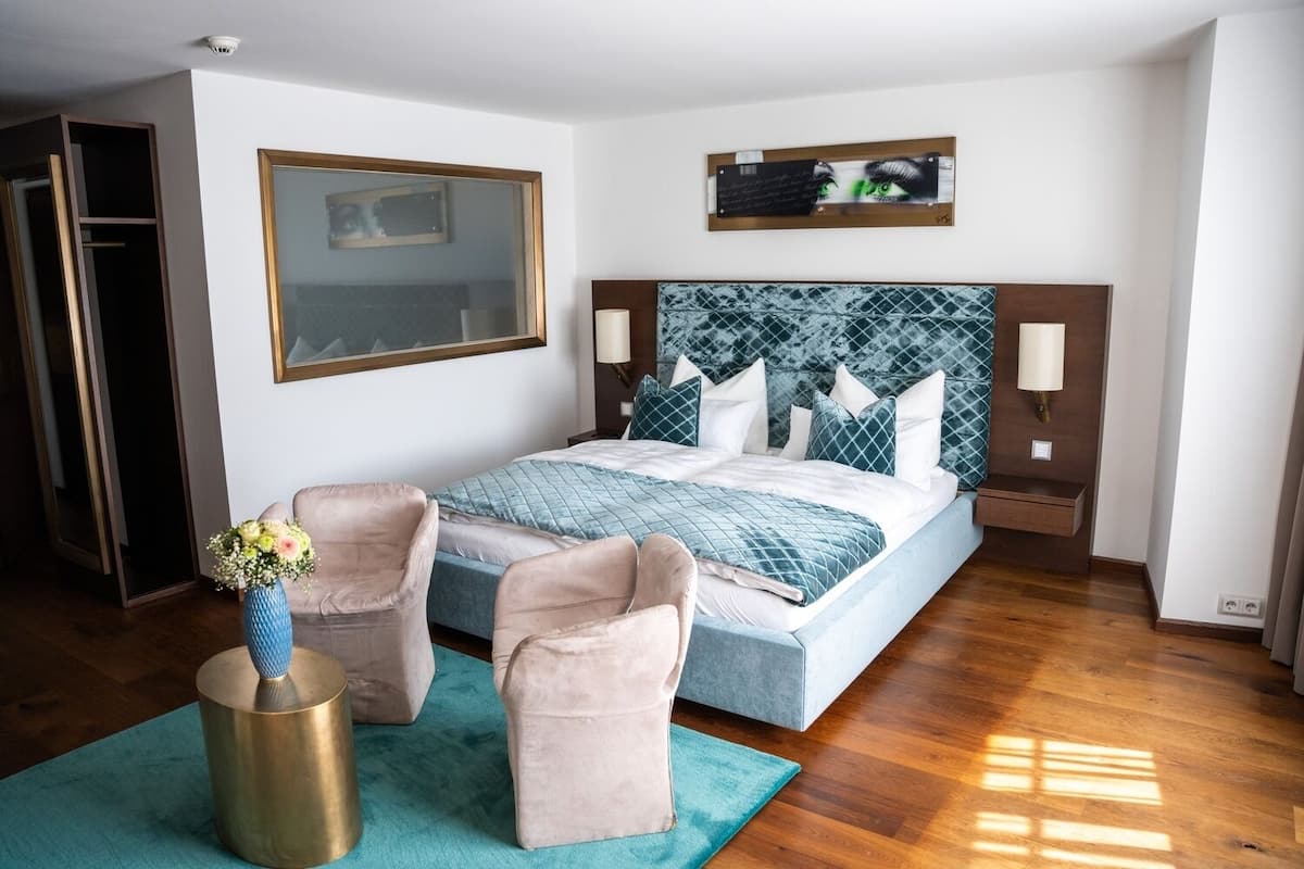 A bedroom with a bed, two chairs, a vase with flowers, a mirror, and a wall-mounted artwork.