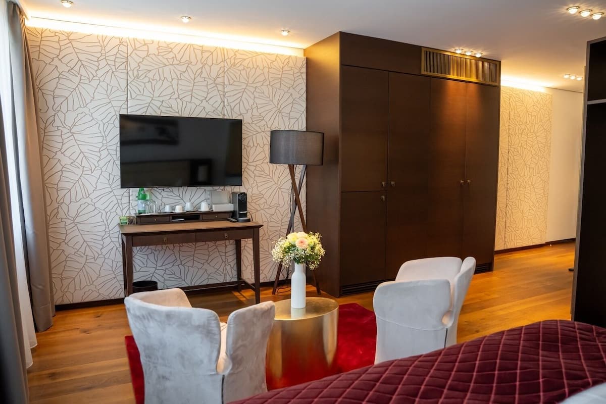 A hotel room with a flat-screen TV, a desk with a lamp, a wardrobe, and two chairs.