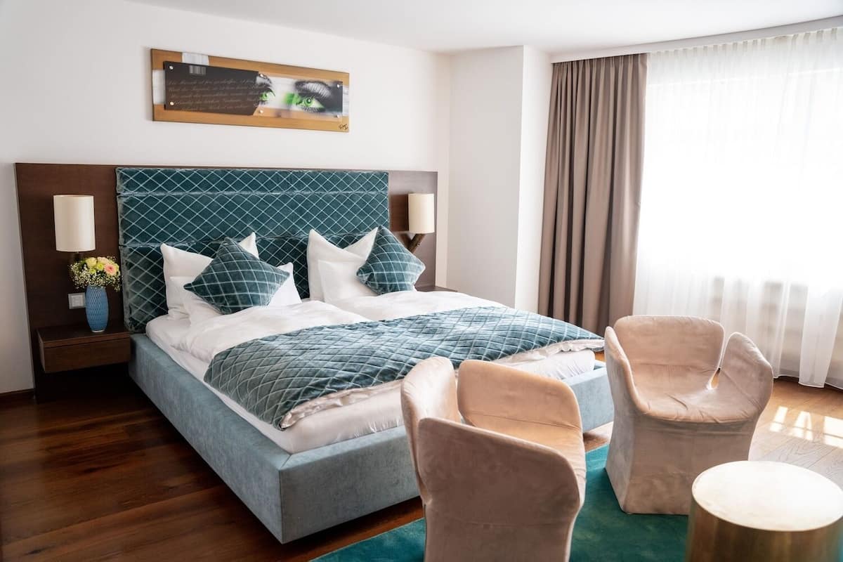 A hotel room with a bed, two chairs, a nightstand with a lamp, a vase with flowers, and a window with curtains.