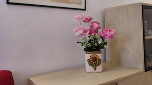 Ein gerahmtes Bild mit Früchten und Blumen an einer Wand, ein Blumentopf mit rosa Blüten auf einem Schrank und ein roter Sessel.