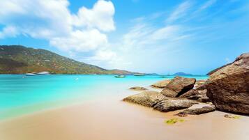 Une plage avec une eau turquoise et limpide, de gros rochers et des bateaux qui se dessinent à l’horizon.
