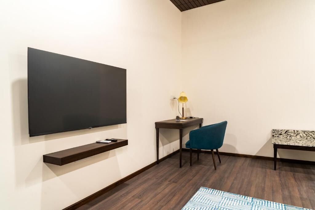 A modern room with a flat-screen TV, a small table with a lamp, a blue chair, and a patterned bench.