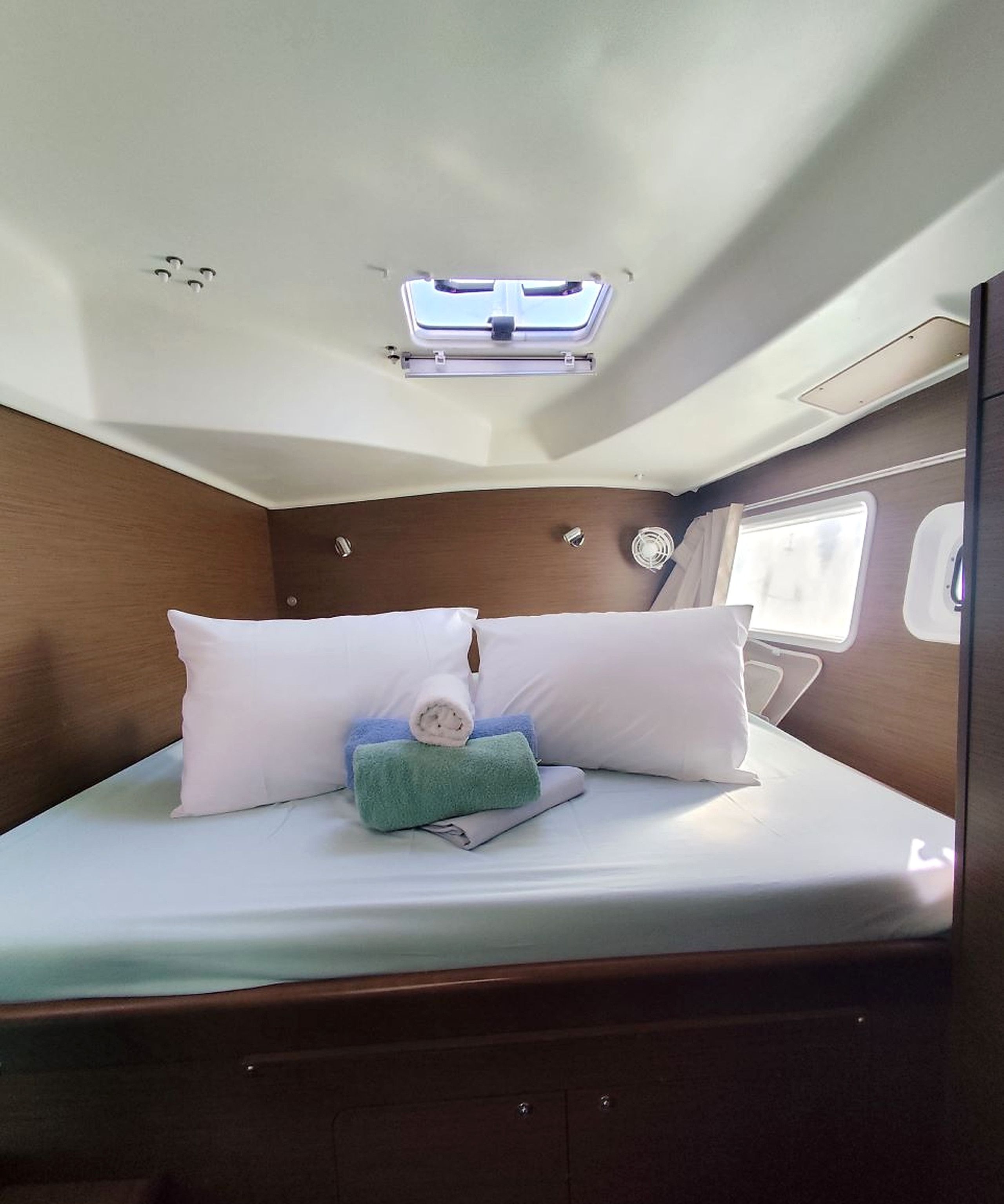 Una cama compacta con ropa blanca, un tragaluz arriba y una ventana pequeña a la derecha.