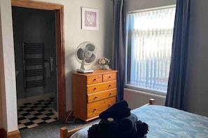 A bedroom with a bed, a wooden dresser, a standing fan, a radiator, a window with curtains, and a checkered floor. - 1 Apartment Sleeps 2 (Sutton-in-Ashfield)