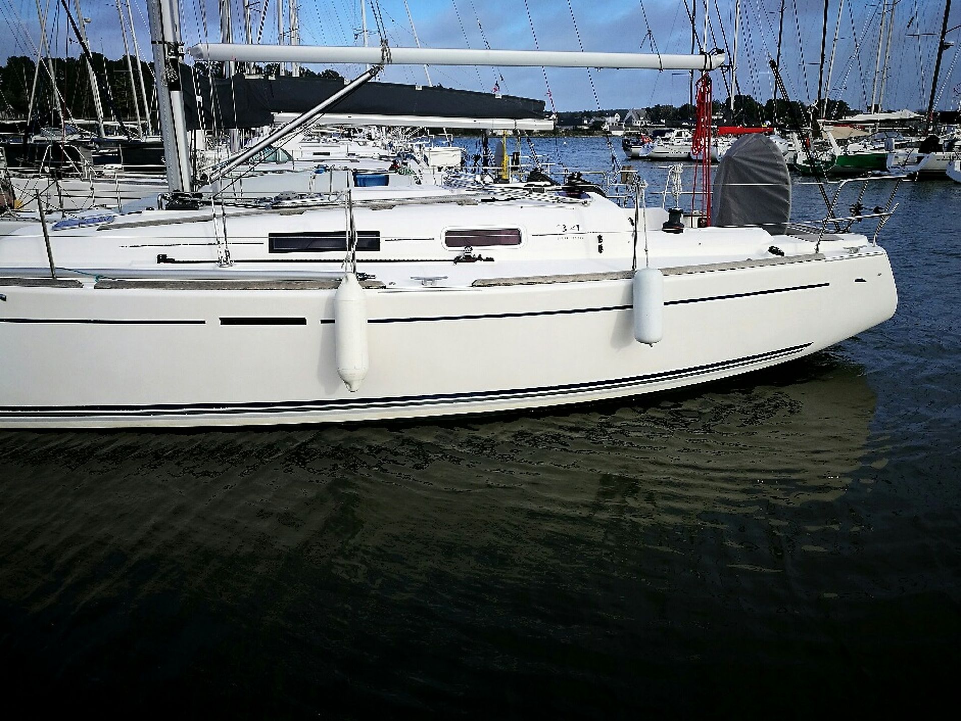 Un voilier blanc est amarré dans une marina, avec d’autres bateaux en arrière-plan.