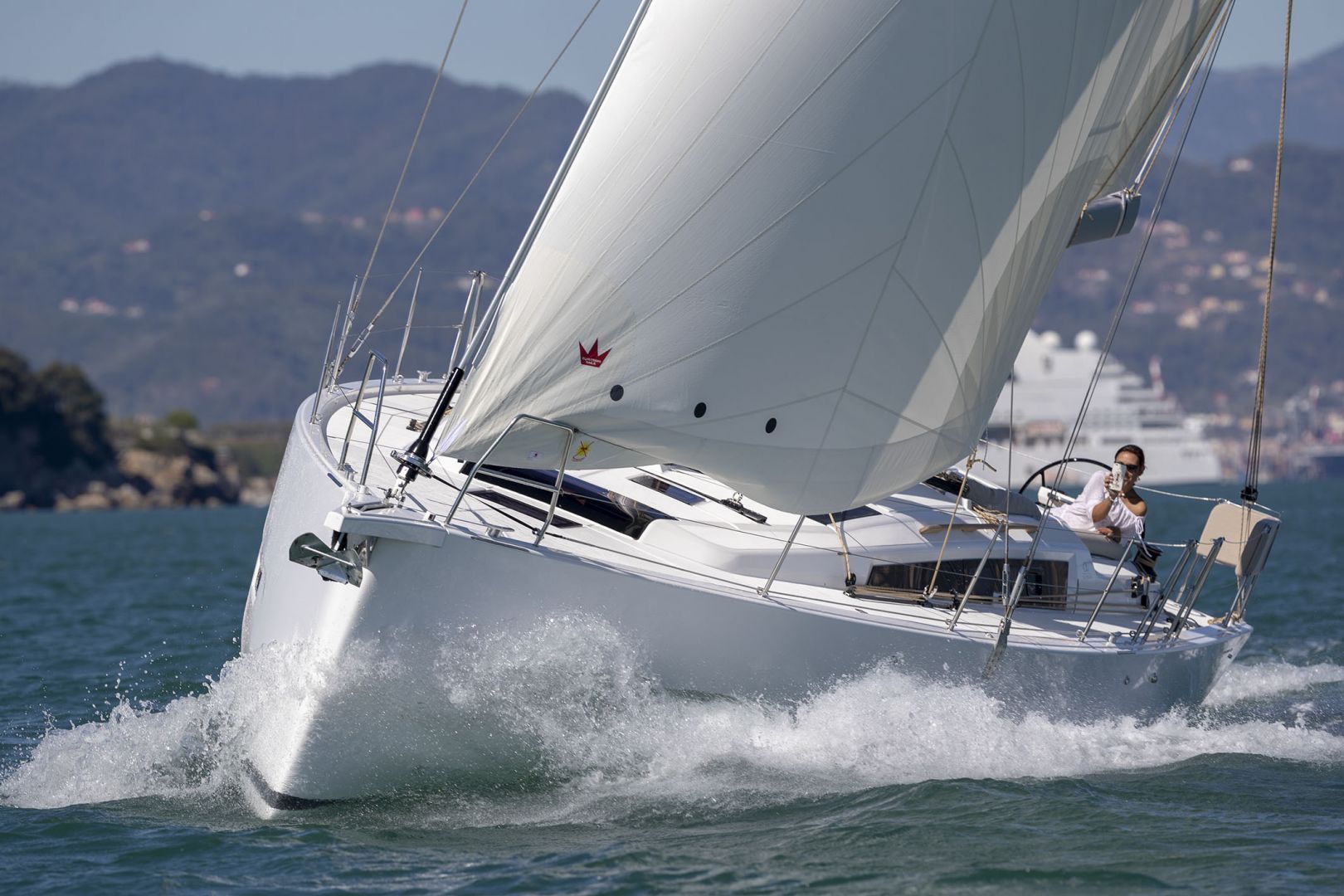 A sailboat with a white hull and a person steering, sailing on water with a mountainous coastline in the background.