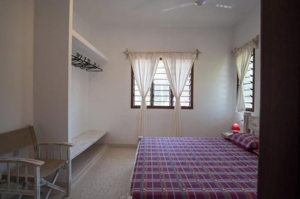 Une chambre à coucher comprenant un lit, un banc, une chaise, une fenêtre avec des rideaux et un ventilateur de plafond.