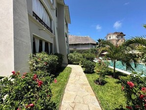 A pathway leading to a pool area with a building on the left and a thatched-roof structure in the background. - Casa Di Philo 2 – Elegant Coastal Escape in Watamu with Pool & Garden (J2XF+F9P, Watamu)