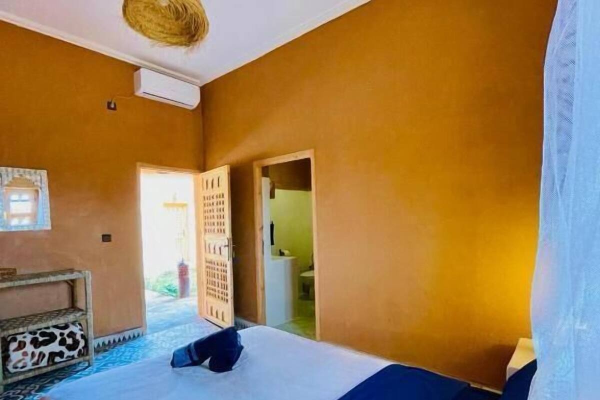 Un dormitorio con cama, lámpara colgante, ventana y puerta que da a otra habitación.