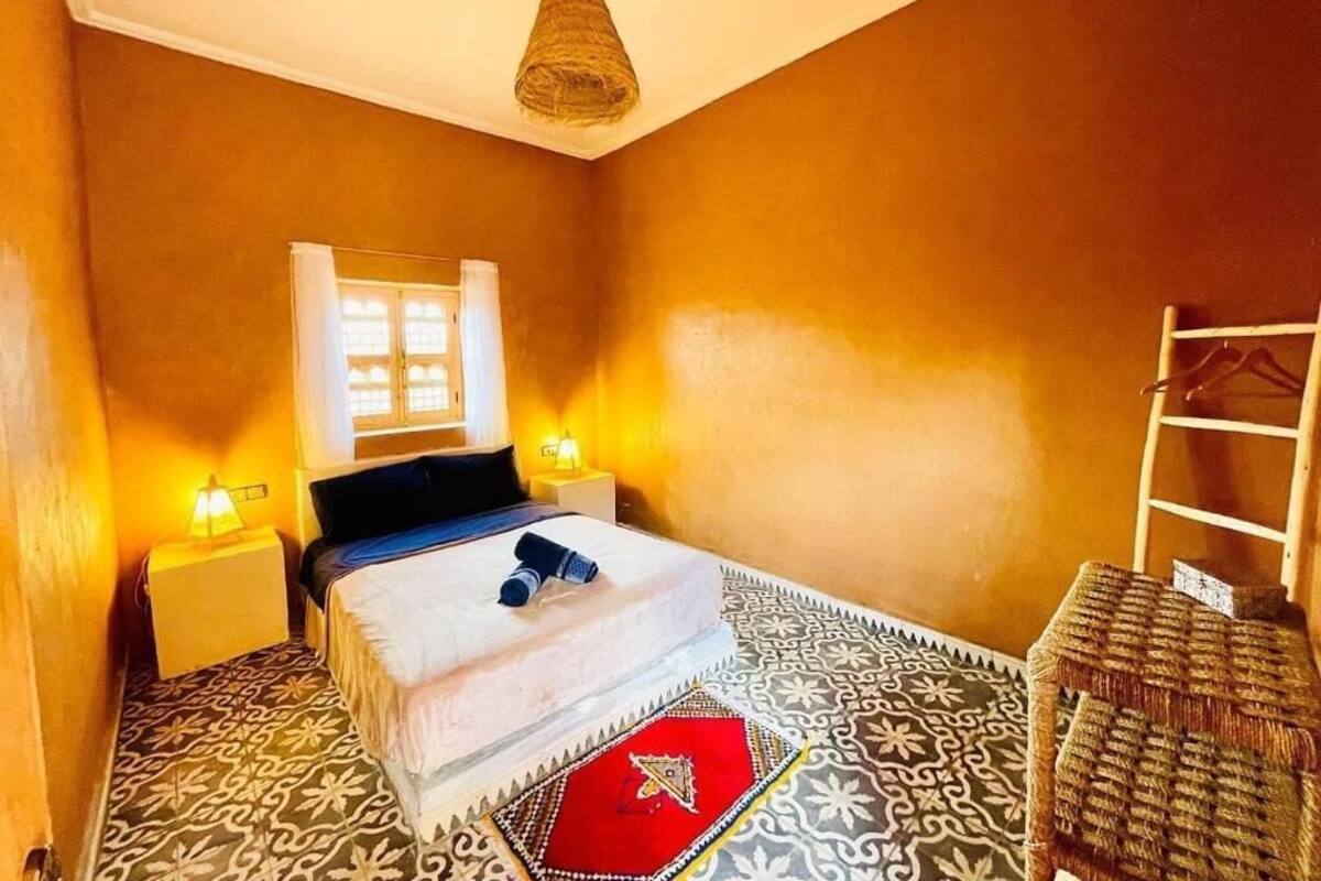 Un dormitorio con una cama, lámparas de noche, un tapete estampado, una silla y una lámpara colgante.