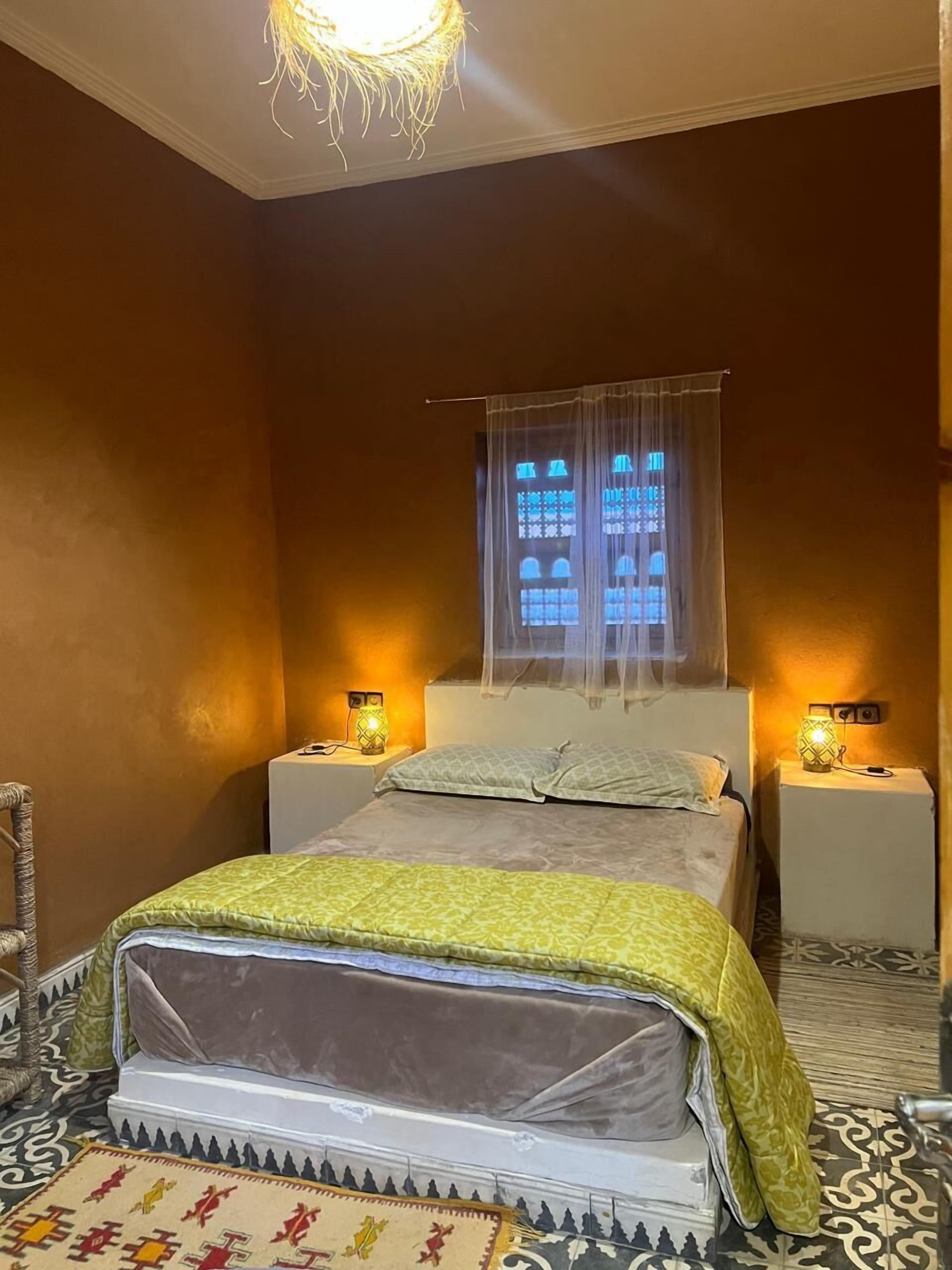 Un dormitorio con una cama, dos mesitas de noche con lámparas, una ventana con cortinas y una alfombra estampada.