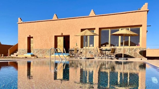 Ein Pool mit klarem Wasser, ein Gebäude mit steinverkleideter Fassade und Sitzgelegenheiten im Freien mit Sonnenschirmen.