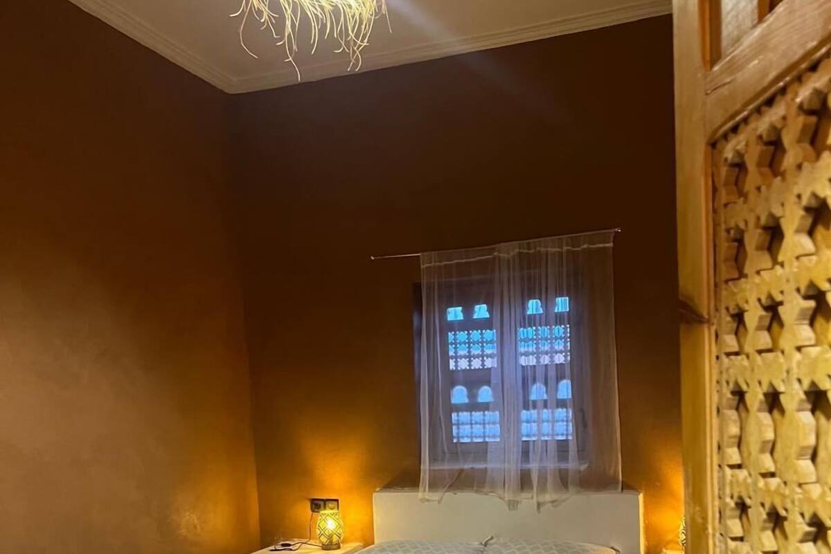 Una habitación con cama, ventana con cortinas, mesita de noche con lámpara y una puerta decorativa.