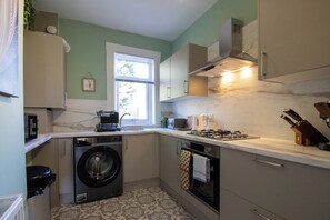 A compact kitchen with a washing machine, oven, and a window with blinds. - Presented by Aurora Short Stays! This spacious 2-bedroom property at Albion St, Motherwell is perfectly located near shops, cafés, and everyday amenities. Sleeping up to 4 guests, it features 1 king bed and 2 single beds for a comfortable stay. (North Lanarkshire)
