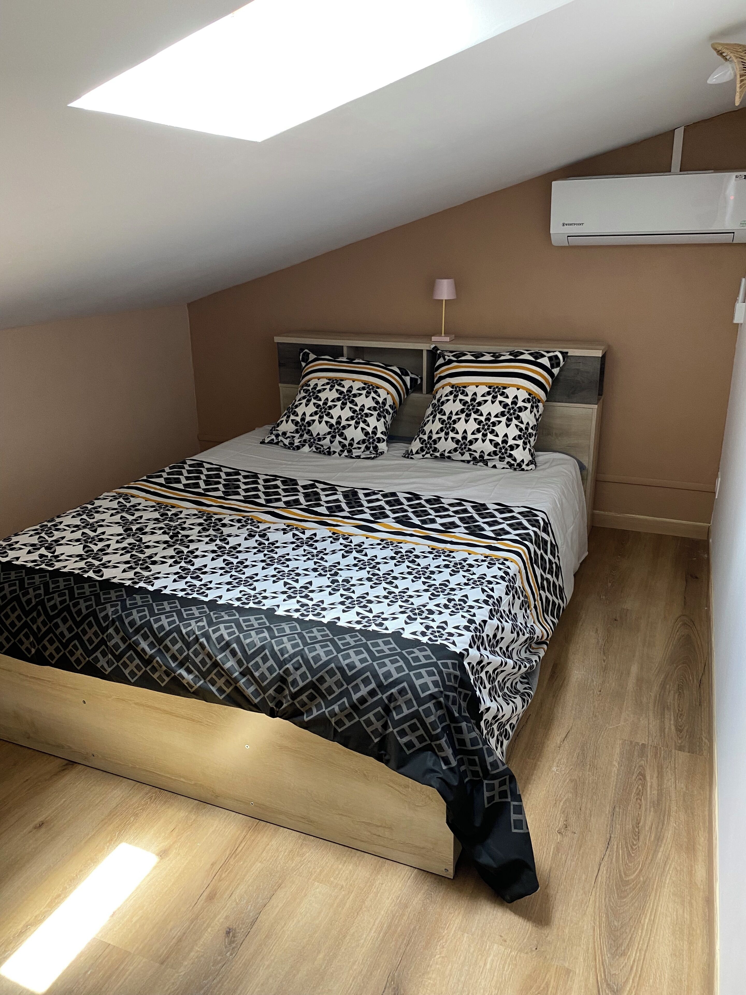 A bedroom with a sloped ceiling, a bed with patterned bedding, a bedside lamp, and an air conditioning unit.