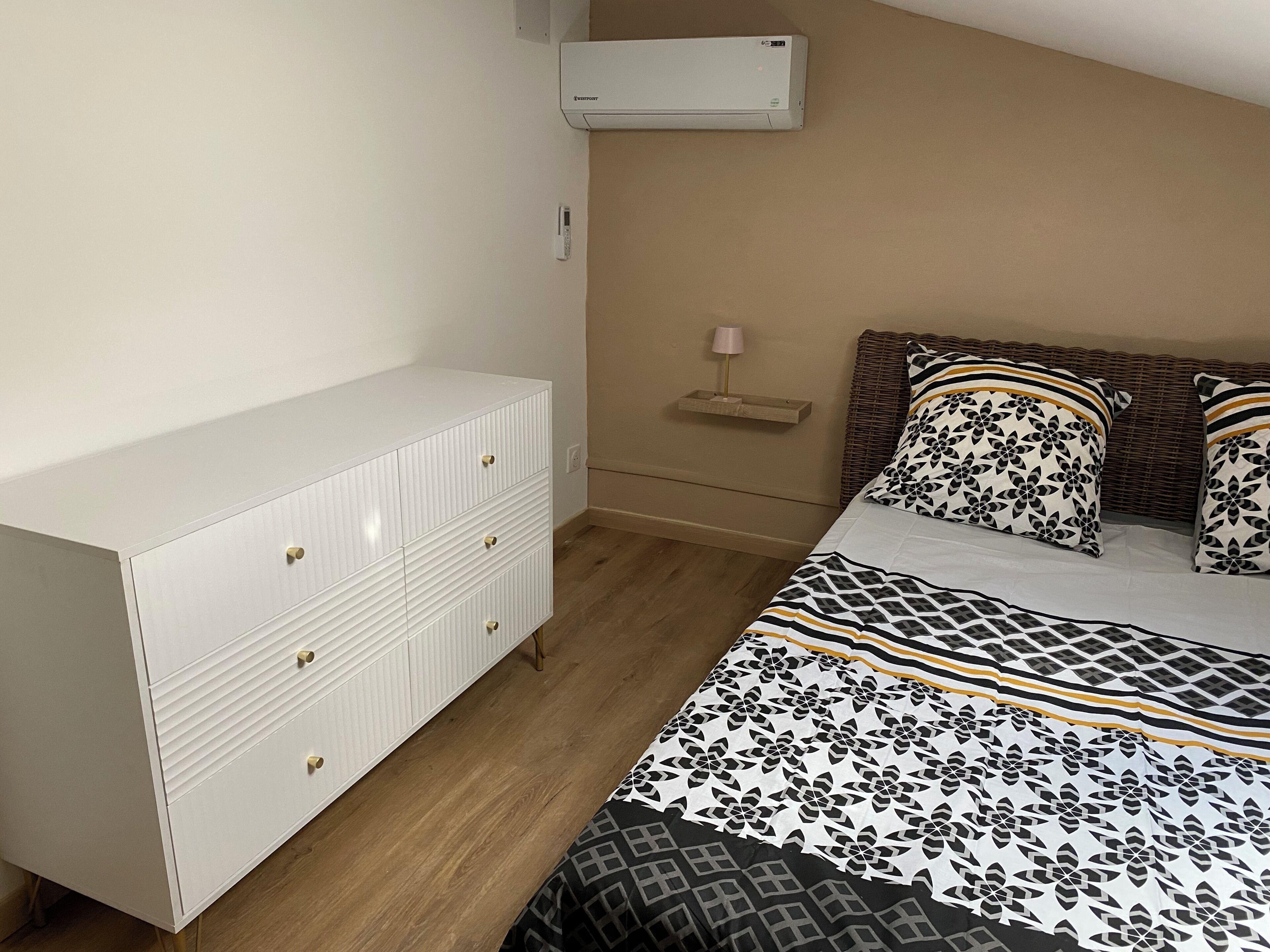 Un dormitorio con una cama, un tocador blanco, una mesita de noche, una lámpara y un equipo de aire acondicionado.