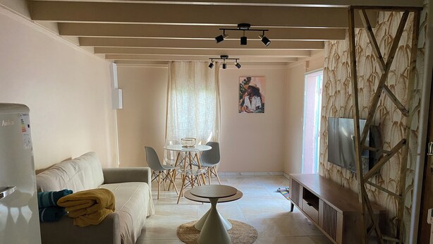 Een woonkamer met een bank, een salontafel, een eettafel met stoelen, een televisie bevestigd aan een behangwand, en een koelkast.