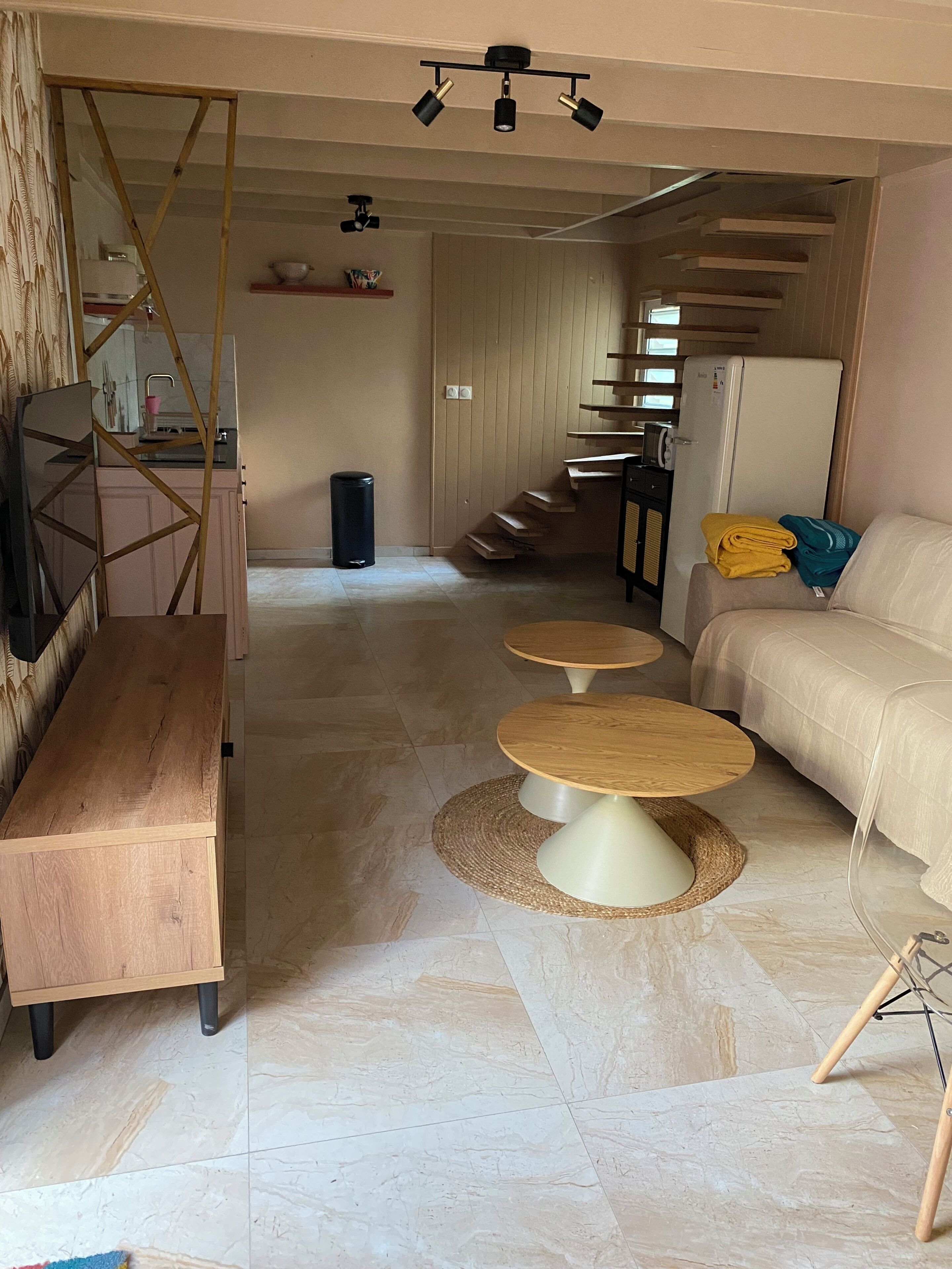 Una sala de estar moderna con un sofá, dos mesas redondas, un banco de madera, una escalera y una zona de cocina.