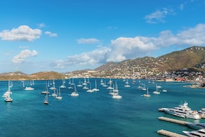 Uma marina com vários veleiros atracados, uma cidade costeira ao fundo e um céu azul claro.