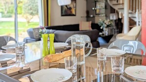 Une table à manger dressée avec des assiettes, des verres et une baguette, donnant sur un salon et un espace extérieur.