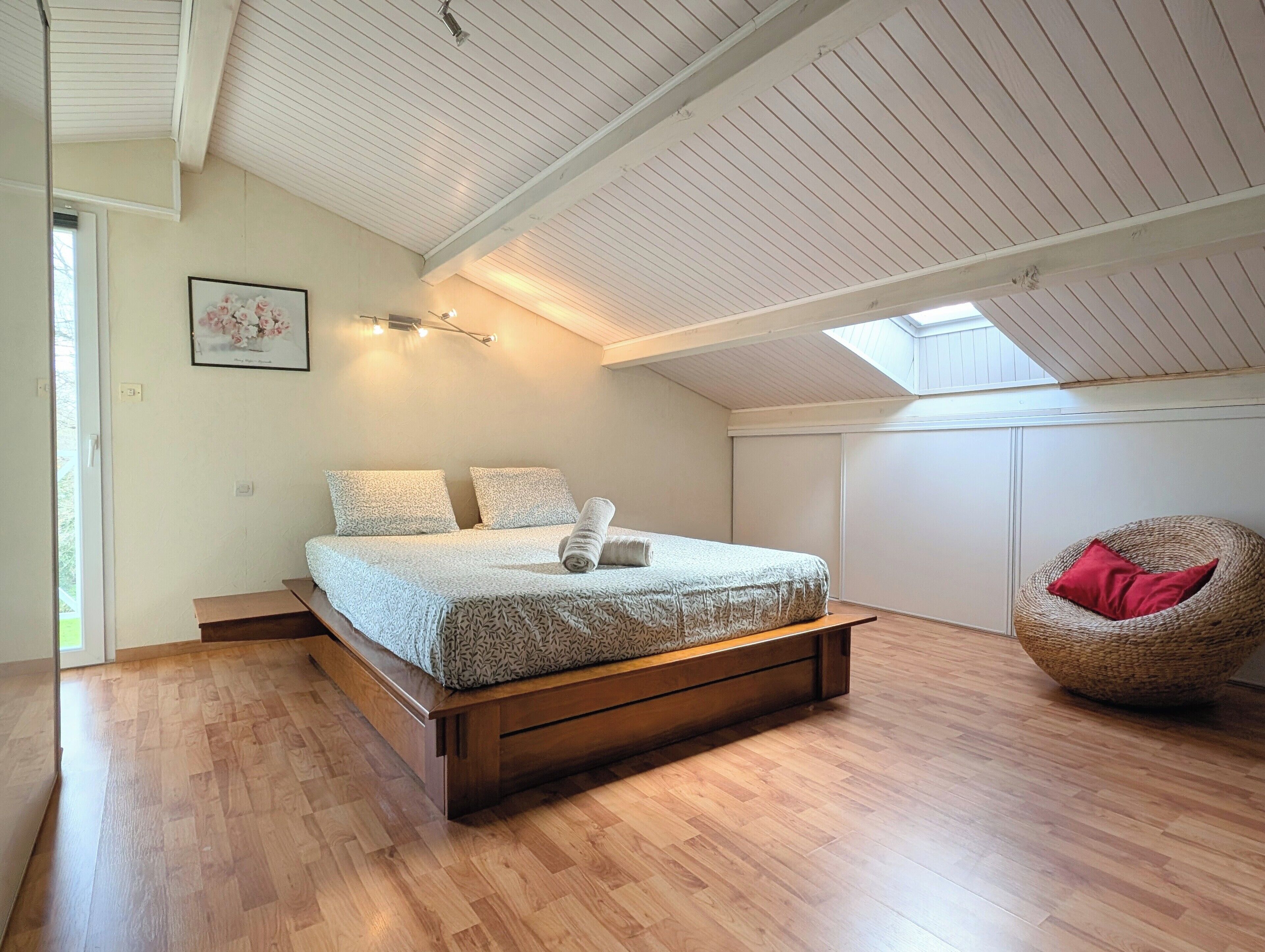 Un dormitorio con techo inclinado, una cama con ropa de cama estampada, una silla y un tragaluz.