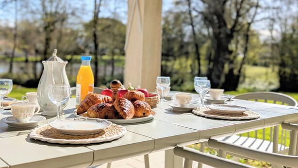 Une table à manger installée à l’extérieur, avec des croissants, une bouteille de jus d’orange et des tasses à thé.
