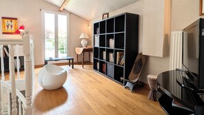 Un salon moderne avec un plancher en bois, un fauteuil blanc, une bibliothèque noire, un téléviseur et une batterie de percussion.