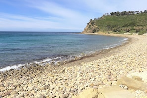 Una playa de guijarros con agua cristalina, una costa rocosa y una colina con árboles al fondo.