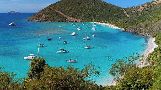 A coastal area with boats anchored in clear turquoise waters, surrounded by lush greenery and a prominent hill.