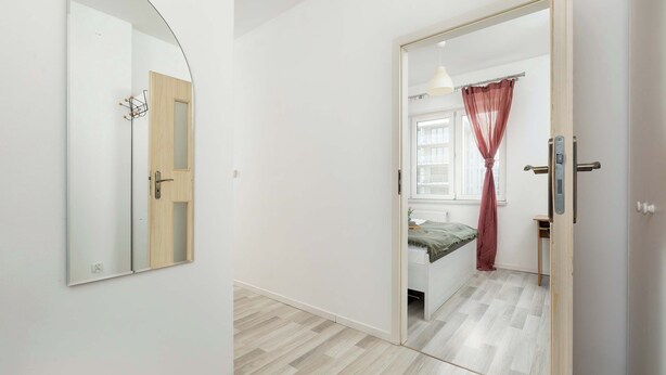 A room with a wooden door, a mirror, a bed with a green blanket, a window with sheer curtains, and a light fixture. - Cozy Apartment | Workspace (Kraków)
