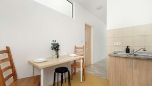 A compact kitchen with a dining area, a wooden chair, a stool, a white plate, a glass, and a bottle on the counter. - Cozy Apartment | Workspace (Kraków)