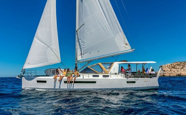 Un velero con varios pasajeros en la cubierta, una zona de camarotes y una costa rocosa al fondo.