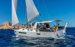Un velero con varios pasajeros, una costa rocosa al fondo y mar en calma.