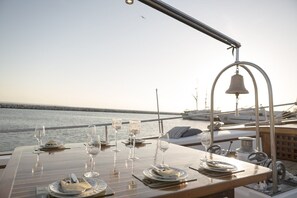 Une table dressée pour un repas, avec des verres et des couverts, une cloche, et une vue sur la mer.