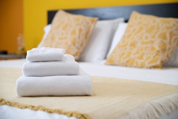 A neatly made bed with a white and beige bedspread, two pillows with a textured pattern, and a stack of folded white towels.