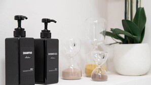 Dos botellas negras con las etiquetas 'SHAMPOO' y 'SHOWER GEL' sobre una superficie blanca, junto a relojes de arena y una planta en maceta.