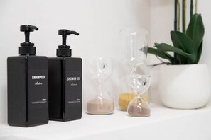 Dos botellas negras con las etiquetas 'SHAMPOO' y 'SHOWER GEL' sobre una superficie blanca, junto a relojes de arena y una planta en maceta.