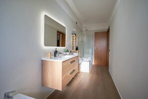 Une salle de bain moderne avec un meuble-lavabo en bois, un grand miroir, une douche et une porte donnant sur une autre pièce.
