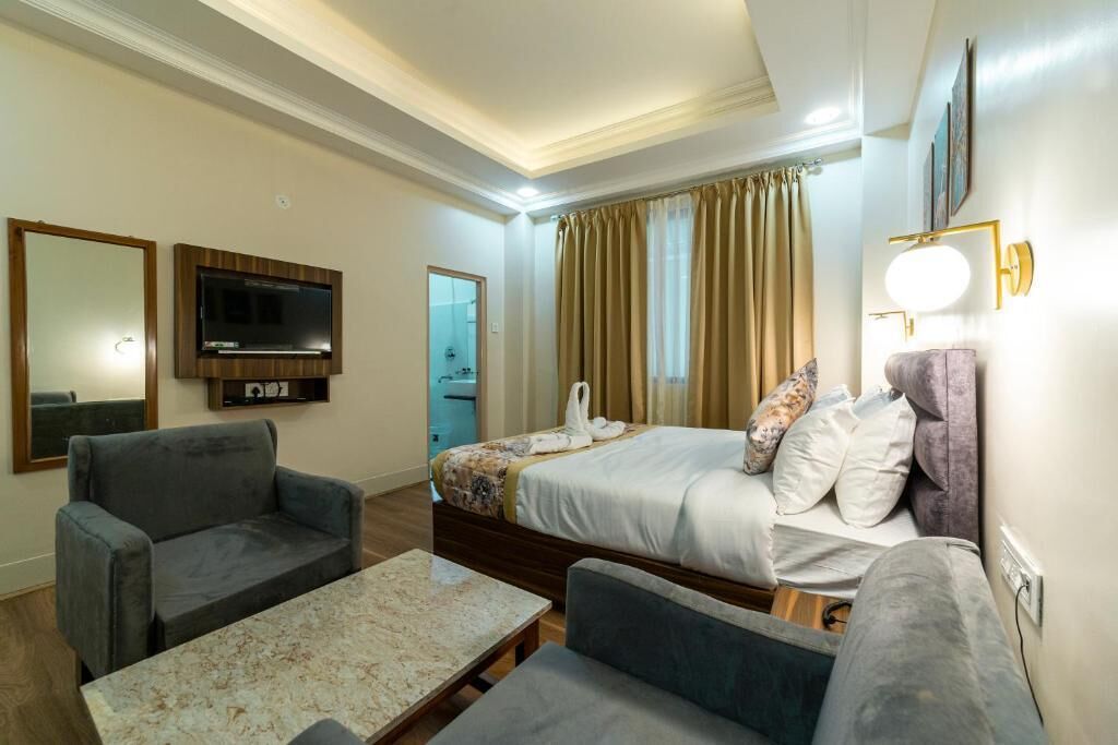 A hotel room with a bed, two armchairs, a small table, a TV, and a bathroom visible through a door.